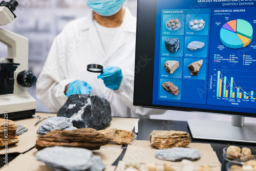 Fotografie Geologist examines rock samples with a hand lens beside a microscope and computer screen mineral analysis charts in a modern laboratory