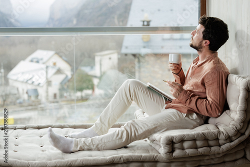 Man with coffee and notebook looking out through a large window at a village