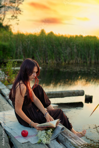 Fotografie Young woman reading a book on a wooden pier by the lake during a peaceful golden