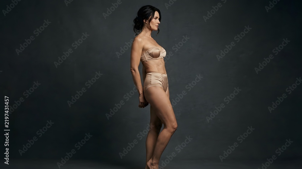 Obraz premium Side profile of a fit woman in lingerie posing against a dark background.