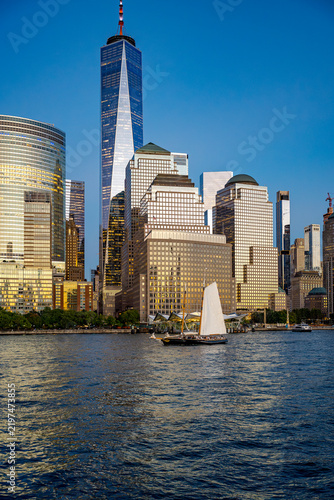 Sailboat cruising Hudson River past One World Trade Center in New York City. A white sailboat sails on blue river water in front of One World Trade Center and financial district skyscrapers.