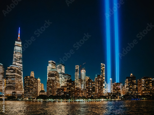 Tribute in Light beams behind Lower Manhattan skyline at night in New York City. Two bright vertical blue light beams shine into night sky behind illuminated skyscrapers of Manhattan along water.