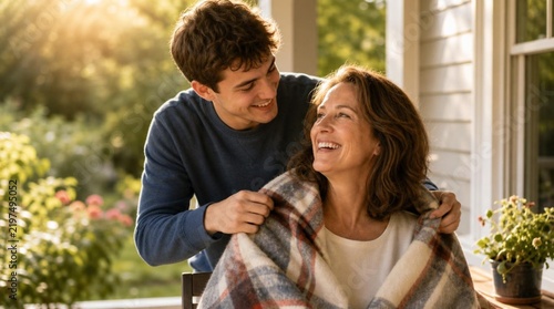 Young man wrapping a soft plaid blanket around the shoulders of his smiling mother on a sunlit porch, sharing a warm, joyful family moment together outdoors in golden sunlight