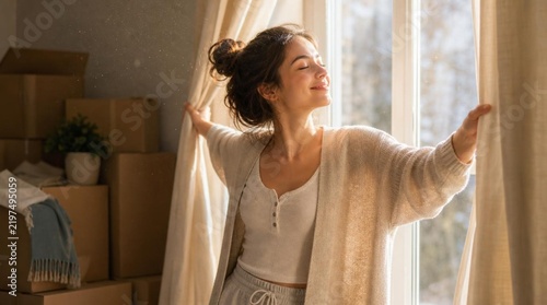 Young woman in casual loungewear joyfully parting beige curtains at sunlit window of her new apartment, eyes closed, surrounded by moving boxes
