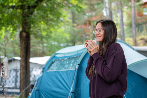 Young woman drinking coffee from a mug while camping in a forest tent site