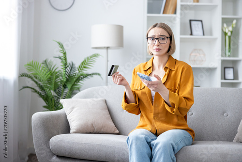 Фотография Financial stress and online shopping problems, worried woman using a smartphone and credit card for payment on a sofa in a home living room