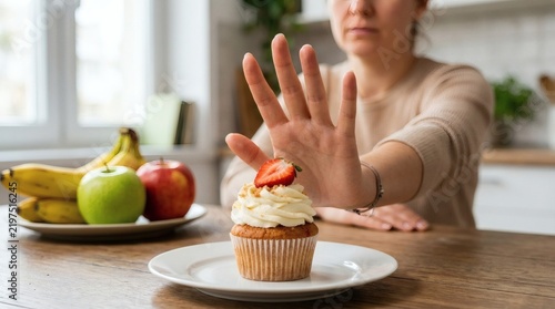 Person gesturing stop with hand towards cupcake topped with strawberry while sitting at kitchen table with fruit platter featuring bananas and apples.