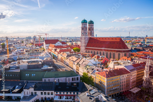 A panoramic view of Munich featuring the iconic Frauenkirche with its twin green-domed towers at sunset, surrounded by historic rooftops and the cityscape under a bright blue sky.