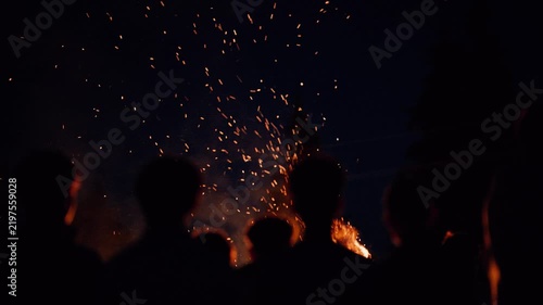 People gather around a fire at night, watching sparks rise in the air