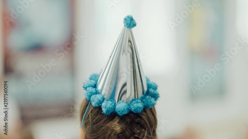 Child wears a party hat during a celebration in a colorful indoor space with decorations and people in the background