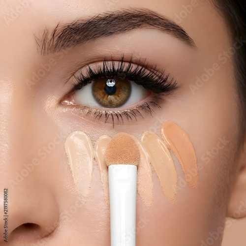 Extreme close-up of a woman's eye and cheek featuring several swatches of liquid foundation in various skin tones being blended with a professional makeup sponge applicator tool