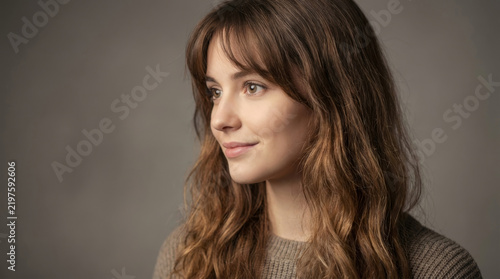 Woman with long wavy hair gazes thoughtfully in a close portrait. Soft studio light shapes her face against a neutral background. Beauty and lifestyle concept.