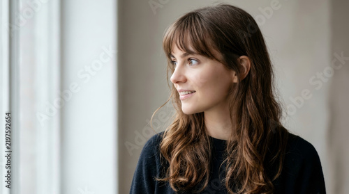 Young woman gazes thoughtfully toward a bright window. Soft daylight illuminates her face in a quiet home interior. Portrait concept for reflection, hope, and wellbeing.