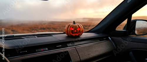 Glowing jack-o’-lantern with a scary face sits on a car dashboard at sunset over a field. Great for Halloween auto ads, festive banners, event posters, dealership promos, and social media.