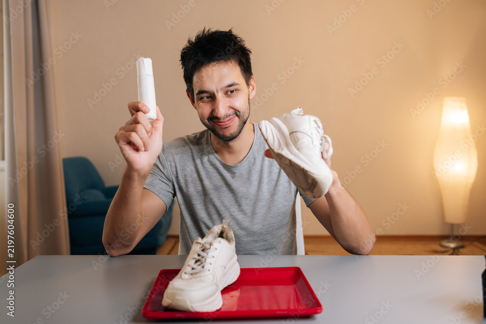 Obraz premium Vertical portrait of cheerful young man holding white sneaker and shoe cleaner bottle, preparing to clean footwear at home, demonstrating process of refreshing shoes and maintaining appearance.