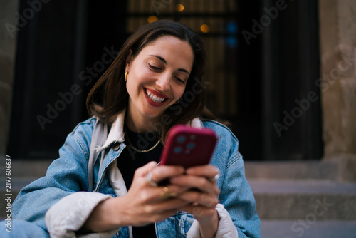 Radiant Caucasian female student laughing while looking down at smart phone sitting on steps, enjoying cybersecurity safe social messaging and dynamic modern digital connection