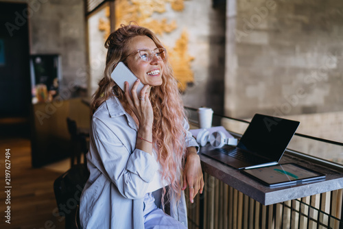 Caucasian female student in glasses laughing during mobile phone call near open laptop and tablet, discussing remote e-learning course assignment in modern lounge workspace studio.