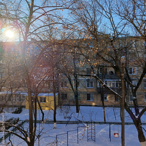 A winter courtyard covered in snow. Trees with remaining leaves, a playground, and a multi-story building are all illuminated by the bright morning sun. Beauty in nature.