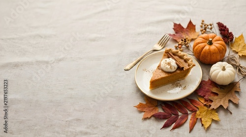 A slice of homemade pumpkin pie with whipped cream and cinnamon stick on a white plate with autumn leaves and small gourds for Thanksgiving dinner celebration.