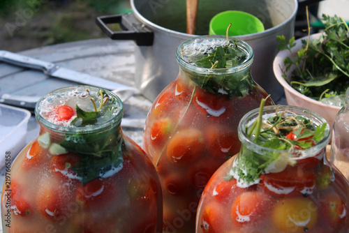 Fotografie Three glass jars filled with fresh cherry tomatoes and aromatic herbs in brine for home pickling, showcasing traditional food preservation outdoors in natural light