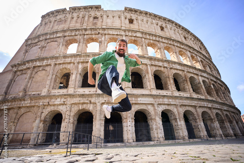 Happy Caucasian millennial tourist jumping energetically in front of the Colosseum in Rome. Wide-angle travel scene conveying excitement, freedom and adventure at an iconic landmark.