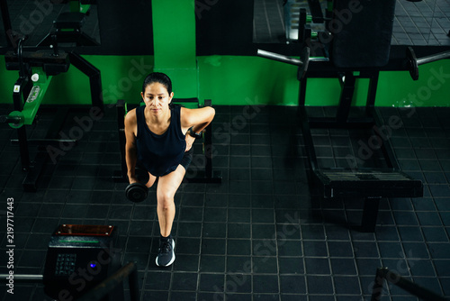 Fotografie A woman completes a weighted lunge while moving across a gym floor, holding dumbbells and maintaining controlled form