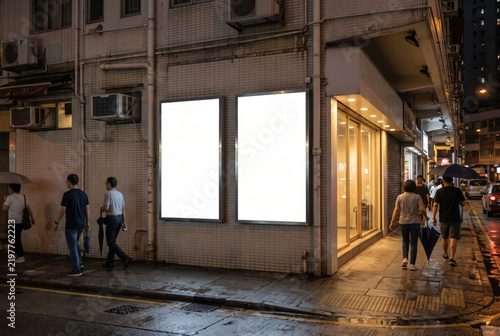 Two blank white advertising lightboxes on a tiled building wall on a wet city street at night, with pedestrians walking past under warm light.