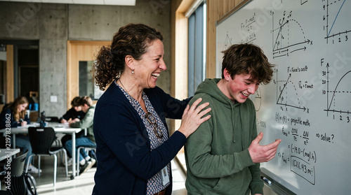 Smiling teacher and male student laughing together in a modern classroom near a whiteboard covered in mathematical formulas