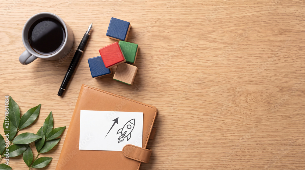 Fototapeta premium Business Startup Launch Concept with Rocket Drawing on Notebook Beside Coffee Cup Pen and Wooden Blocks on Wood Table Background for Success Strategy Planning and Creative Growth Vision Top View