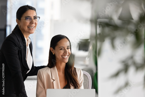 Happy diverse businesswomen laughing during meeting in corporate office, representing effective teamwork completion, mentor and apprentice friendly relations, joint business achievements and success