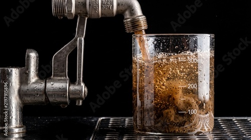 A heavy, functional stainless steel utility pipe dispensing an authentic, turbulent rush of murky, rust-brown, sediment-heavy tap water directly into a clean glass