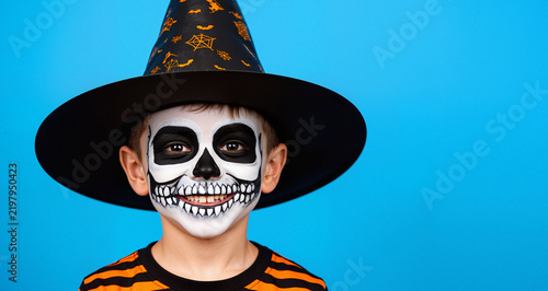 Smiling boy in skeleton face paint with witch hat, Halloween child with skull makeup on blue background, Fun Halloween portrait with copy space
