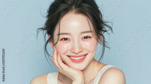 Beautiful smiling young Asian woman with radiant glowing skin, perfect teeth, clean natural makeup, and hand on chin over blue background for beauty skincare concept
