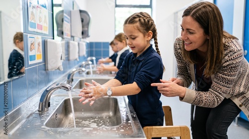 Teacher helping young girl wash hands in school bathroom