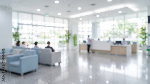 Blurred background image of a modern hospital lobby or clinic reception area featuring waiting patients sitting on chairs, reception desk with staff, large windows, and shiny tiled floors in bright.