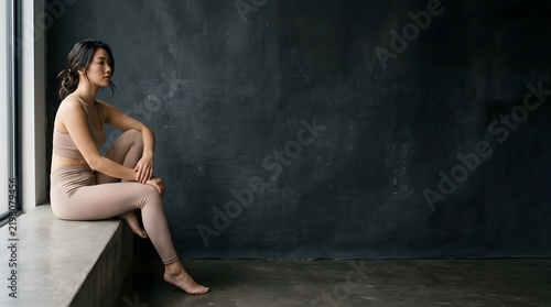 A contemplative woman sitting on a windowsill inside a dark room.