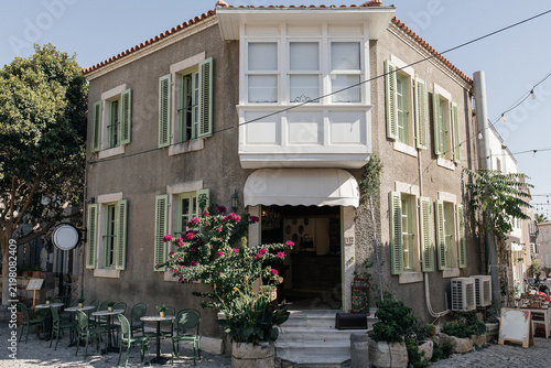 Aegean house with green shutters and bay window, Alaçatı, Turkey. Charming cafe entrance in old Aegean house: stone walls, pastel shutters, terracotta roof tiles, potted plants and green bistro chairs