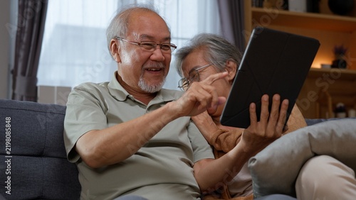 фотографія Senior Asian couple use a tablet at home as the husband points at the screen while his wife watches, representing digital learning, shared technology, online discovery and active retirement life