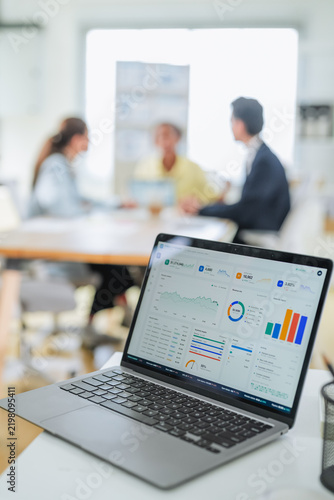 Close-up of a data dashboard on a laptop screen showing sales metrics and performance charts, with a business meeting unfolding softly out of focus behind it.