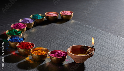 Nine small brass bowls arranged in an arc each holding a different bright coloured powder with a single lit clay oil lamp at the end on a dark slate surface