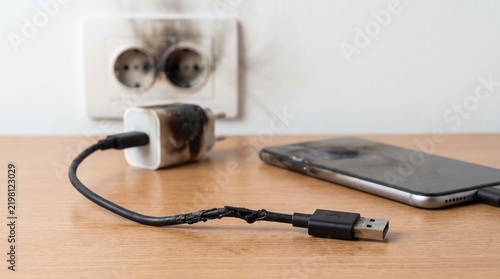 Burnt smartphone charger and damaged power cable plugged into a scorched wall electrical outlet