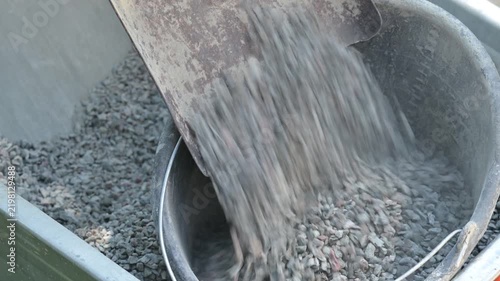 Shoveling crushed stone from a wheelbarrow into a bucket on a construction site. Close-up of gray gravel and dust inside a metal wheelbarrow and an old bucket, material preparation