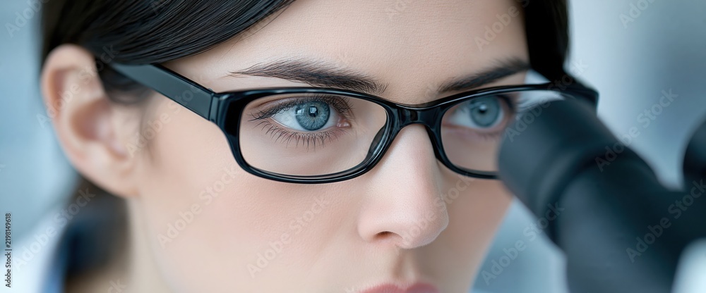Fototapeta premium A young female scientist wearing glasses looks through a microscope in a laboratory setting