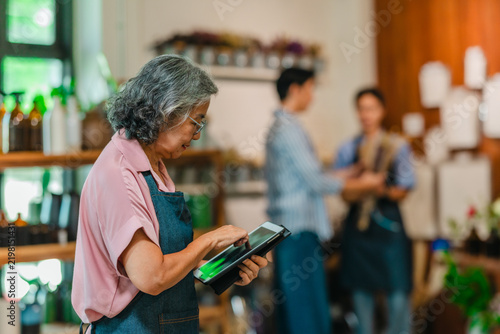 Senior woman shop owner using digital tablet while staff serves customer behind