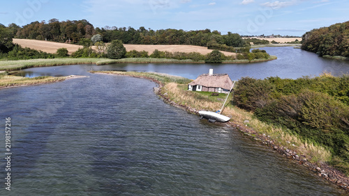 Canvastavla Drohnenaufnahme zeigt ein idyllisches Reetdachhaus mit gestrandetem Segelboot auf einer Landzunge umgeben von blauem Wasser und hügeliger Küstenlandschaft