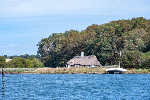 Canvastavla Reetgedecktes Kate-Haus, halb verfallen steht idyllisch an dicht bewaldetem Ufer mit gestrandetem Segelboot am blauen Gewässer unter sonnigem Himmel