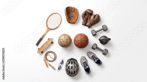 Circular flat lay layout of various sports equipment including wooden racket, skateboard, jump rope, goggles, helmet, sneakers, dumbbells and etc.