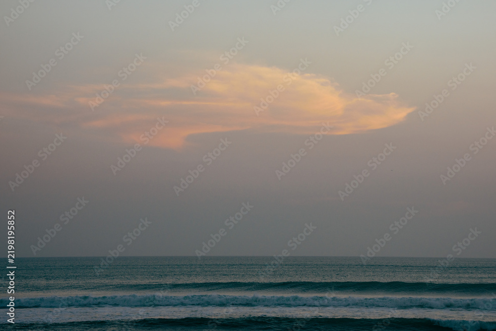 Fototapeta premium A serene sunset over the ocean in Bali features a large, orange cloud against a pastel sky, with gentle waves lapping at the shore.
