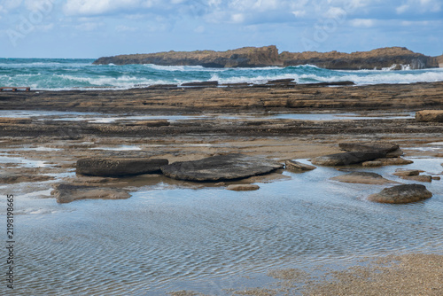 Quadro su tela A rocky coastline with tidal pools and shallow water near breaking ocean waves under a cloudy sky