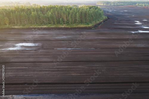 Aerial drone view of a vast industrial peat extraction field bordering a pine forest landscape at sunrise in Estonia.